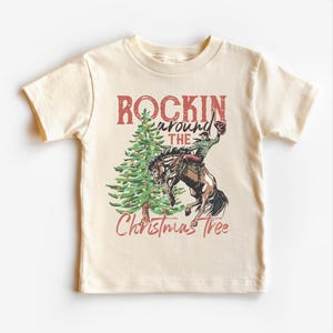 May include: Cream-colored short-sleeved t-shirt featuring a graphic of a cowboy riding a bucking horse next to a Christmas tree. The text reads "Rockin' around the Christmas Tree" in red and green.