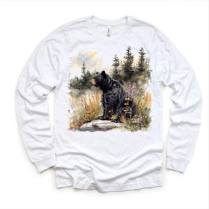 May include: White long-sleeve shirt featuring a watercolor illustration of a black bear sitting on a rock in a forest setting. The bear is surrounded by wildflowers and tall grass. The background includes trees and a cloudy sky.