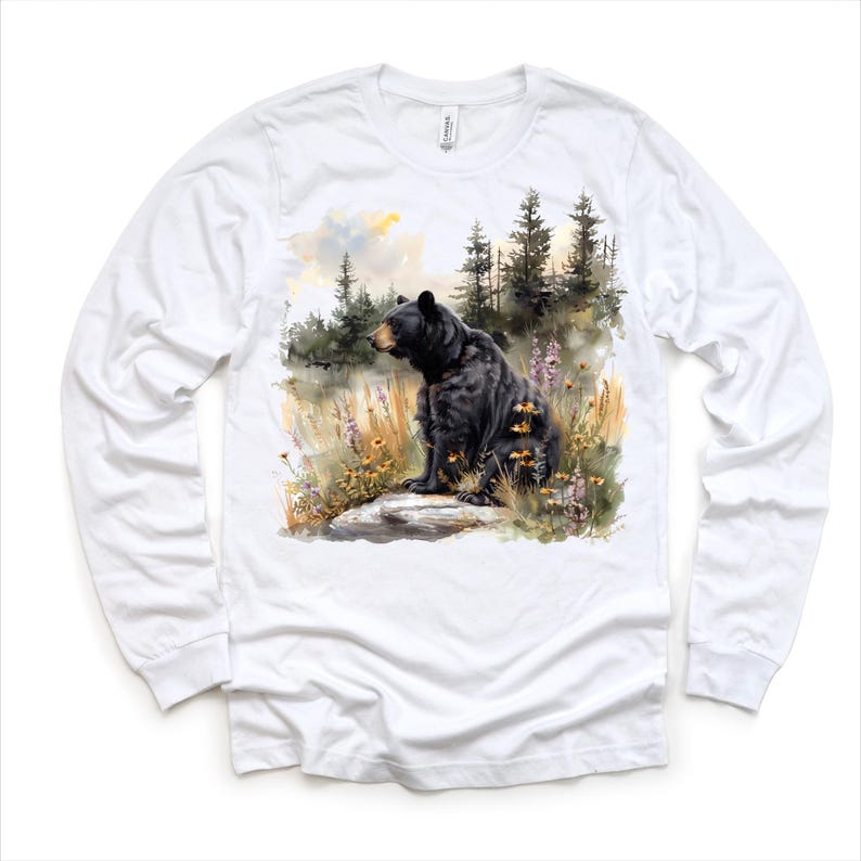 May include: White long-sleeve shirt featuring a watercolor illustration of a black bear sitting on a rock in a forest setting. The bear is surrounded by wildflowers and tall grass. The background includes trees and a cloudy sky.