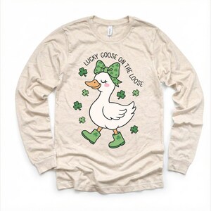 May include: Beige long-sleeve shirt featuring a cartoon goose with a green bow, boots, and shamrocks. The text "LUCKY GOOSE ON THE LOOSE" is printed above the goose. The shirt is made of a soft, comfortable material.