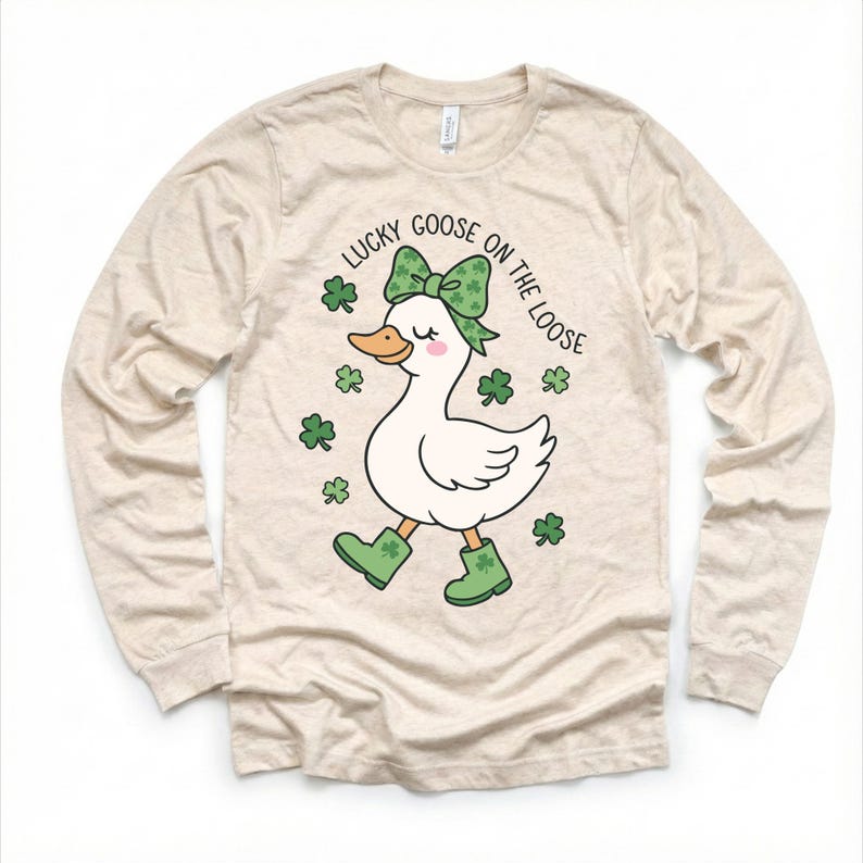 May include: Beige long-sleeve shirt featuring a cartoon goose with a green bow, boots, and shamrocks. The text "LUCKY GOOSE ON THE LOOSE" is printed above the goose. The shirt is made of a soft, comfortable material.