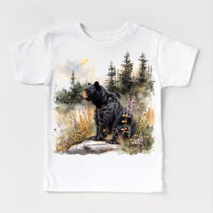 May include: White t-shirt featuring a watercolor illustration of a black bear sitting on a rock in a forest setting. The bear is the focal point, with detailed fur and a naturalistic pose. The background includes trees, wildflowers, and a soft, colorful sky.