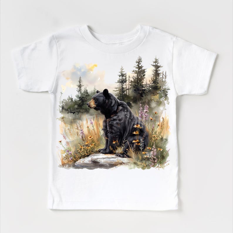 May include: White t-shirt featuring a watercolor illustration of a black bear sitting on a rock in a forest setting. The bear is the focal point, with detailed fur and a naturalistic pose. The background includes trees, wildflowers, and a soft, colorful sky.
