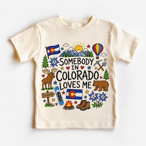 May include: Cream-colored t-shirt featuring a colorful graphic with the text "SOMEBODY IN COLORADO LOVES ME." The design includes a Colorado flag, mountains, a moose, a bear, a hot air balloon, and other outdoor elements.