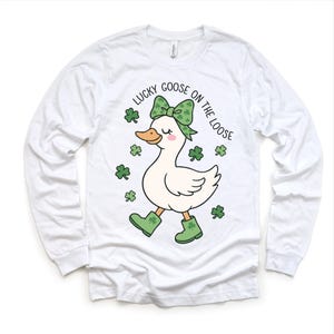 May include: White long-sleeve shirt featuring a cartoon goose with a green bow, boots, and shamrocks. The text "LUCKY GOOSE ON THE LOOSE" is printed above the goose. The shirt is made of a soft material.