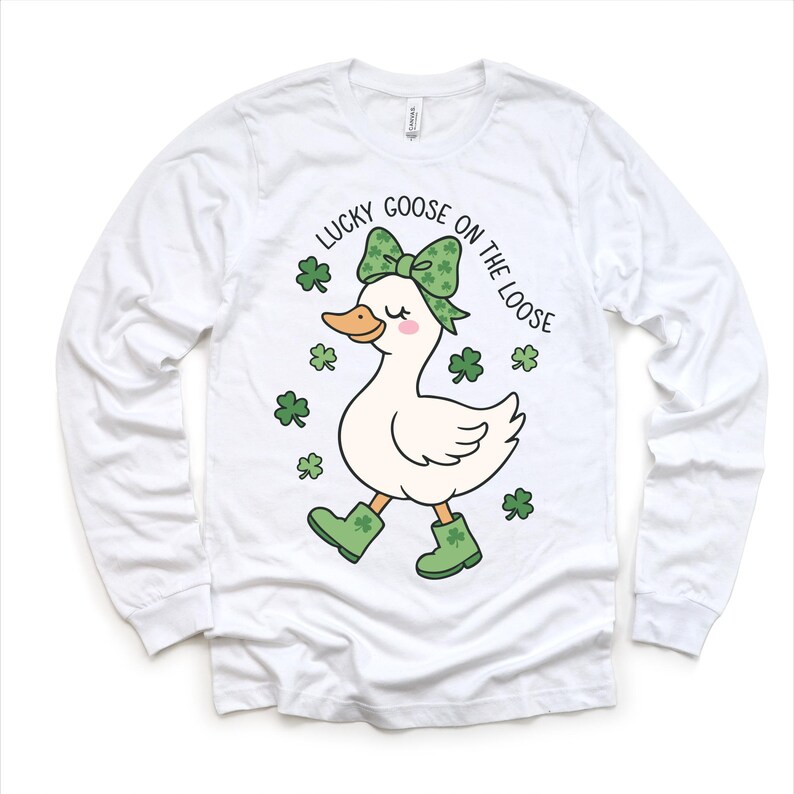 May include: White long-sleeve shirt featuring a cartoon goose with a green bow, boots, and shamrocks. The text "LUCKY GOOSE ON THE LOOSE" is printed above the goose. The shirt is made of a soft material.