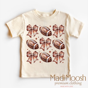 May include: A beige toddler t-shirt with a brown and white football and brown bow pattern. The shirt has the text "Madi Moosh premium clothing made with love" at the bottom.