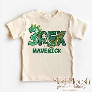 May include: A light beige t-shirt with a green dinosaur graphic that says "3Rex" and the name "Maverick" underneath.