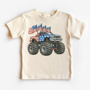 Pode incluir: T-shirt creme com um design de monster truck. O caminh&atilde;o &eacute; decorado com as cores vermelho, branco e azul, e bandeiras americanas. O caminh&atilde;o tem pneus grandes e &eacute; um design patri&oacute;tico.