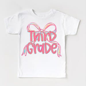 May include: White t-shirt featuring a colorful bow design with the words "Third Grade" in a playful font. The text and bow are in shades of pink, blue, and yellow, creating a cheerful and eye-catching look. A perfect gift for a child.