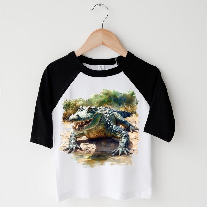 K&ouml;nnte beinhalten: Ein wei&szlig;-schwarzes Baseball-T-Shirt mit einem gr&uuml;nen Alligator-Motiv auf der Vorderseite. Der Alligator ist im Aquarell-Stil gehalten und von gr&uuml;nem Laub umgeben.