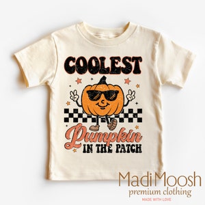 May include: Cream-colored t-shirt featuring a cartoon pumpkin wearing sunglasses and making a peace sign. The text reads "COOLEST PUMPKIN IN THE PATCH" in orange and black. Stars and a checkered pattern are also included.