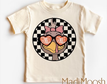 May include: Cream-colored toddler t-shirt featuring a cartoon turkey wearing heart-shaped sunglasses with the words "GOBBLE GOBBLE." The turkey is set against a black and white checkered circle. The shirt has short sleeves.