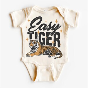 Op de afbeelding: Een cr&egrave;mekleurige baby romper met de tekst "Easy Tiger" in zwart met gouden sterren. Een cartoon tijger ligt op de romper.