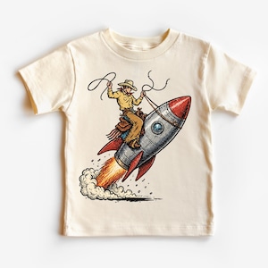 May include: Cream-colored t-shirt featuring a cowboy riding a rocket ship. The illustration shows a cowboy in a hat and chaps holding a lasso, with the rocket emitting flames and smoke. The design is a playful blend of space and Western themes.