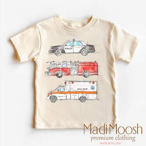May include: A beige short-sleeved t-shirt with a graphic print of a police car, fire truck, and ambulance. The text "POLICE", "AMBULANCE", and "191" are visible on the vehicles. The text "Madi Moosh premium clothing MADE WITH LOVE" is printed on the bottom of the shirt.