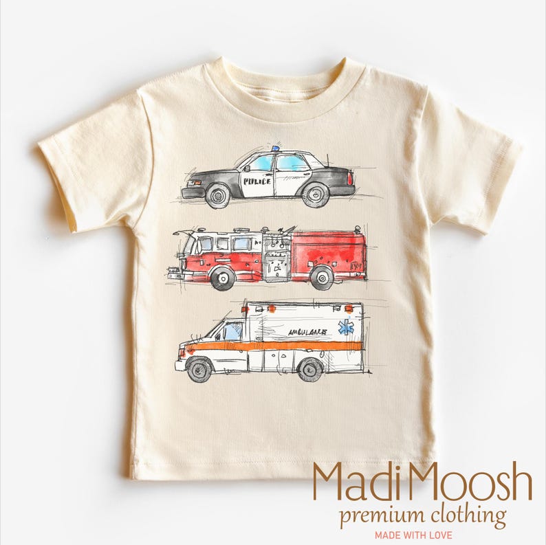 May include: A beige short-sleeved t-shirt with a graphic print of a police car, fire truck, and ambulance. The text "POLICE", "AMBULANCE", and "191" are visible on the vehicles. The text "Madi Moosh premium clothing MADE WITH LOVE" is printed on the bottom of the shirt.