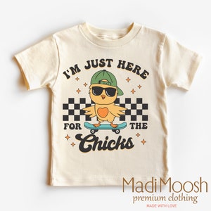 May include: A light beige t-shirt with a cartoon illustration of a yellow chick wearing sunglasses and a green baseball cap, skateboarding. The text "I'm Just Here For The Chicks" is printed on the shirt.