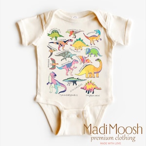 May include: A cream-colored baby onesie with a colorful dinosaur print. The onesie features a variety of dinosaurs, including a Tyrannosaurus Rex, Stegosaurus, and Triceratops. The text "Madi Moosh premium clothing MADE WITH LOVE" is printed on the bottom of the onesie.