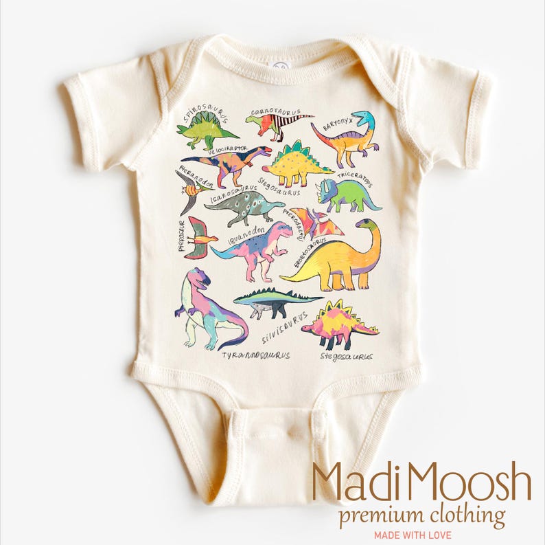 May include: A cream-colored baby onesie with a colorful dinosaur print. The onesie features a variety of dinosaurs, including a Tyrannosaurus Rex, Stegosaurus, and Triceratops. The text "Madi Moosh premium clothing MADE WITH LOVE" is printed on the bottom of the onesie.