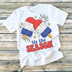May include: White t-shirt with a red, white, and blue popsicle graphic and the text "'tis the season" in red.