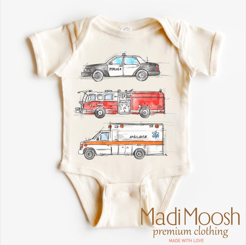 May include: A cream-colored baby onesie with a graphic print of a police car, fire truck, and ambulance. The text "Madi Moosh premium clothing" and "Made with Love" are printed on the bottom of the onesie.