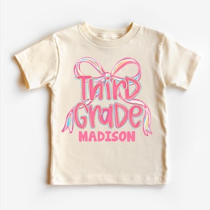 May include: A cream-colored t-shirt featuring a pink bow design with the words "Third Grade" in a script font, and the name "Madison" below. The text is in a gradient of pink, blue, and yellow.