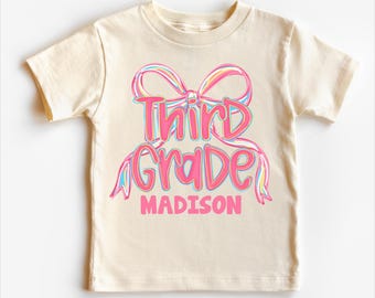 Op de afbeelding: Een crèmekleurig T-shirt met een roze strikdesign en de woorden "Third Grade" in een scriptlettertype, met de naam "Madison" eronder. De tekst is in een verloop van roze, blauw en geel.