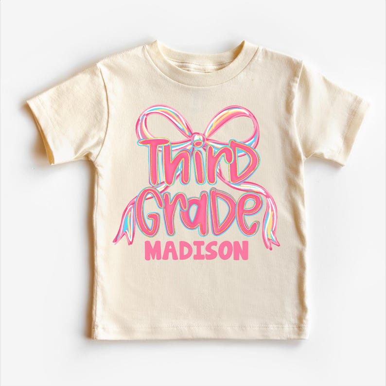May include: A cream-colored t-shirt featuring a pink bow design with the words "Third Grade" in a script font, and the name "Madison" below. The text is in a gradient of pink, blue, and yellow.