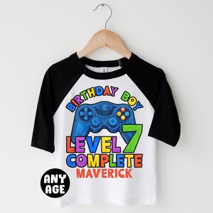 May include: White and black baseball tee shirt with a colorful graphic of a video game controller and the text "Birthday Boy Level 7 Complete Maverick".