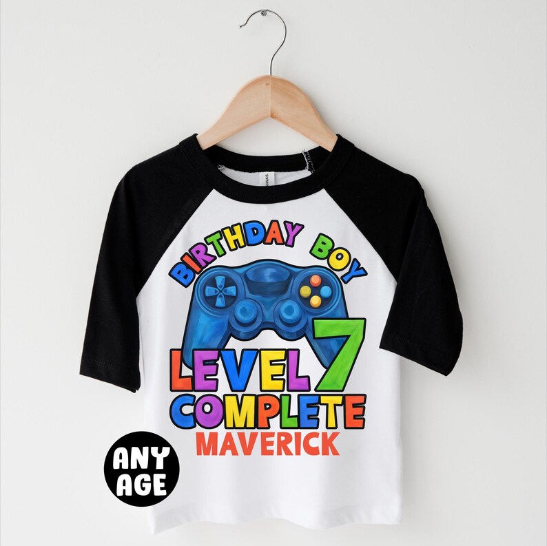 May include: White and black baseball tee shirt with a colorful graphic of a video game controller and the text "Birthday Boy Level 7 Complete Maverick".