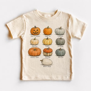 May include: A cream-colored short-sleeve t-shirt featuring a graphic print of ten different pumpkins arranged in rows. Each pumpkin is labeled with its variety name, including Jack-O'-Lantern, Sugar Pie, Cinderella, Baby Boo, Lumina, Jarrahdale, Atlantic Giant, Fairytale, Blue Doll, and White Ghost.