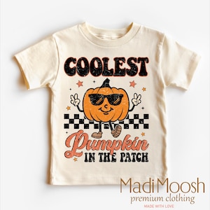 May include: Cream-colored t-shirt featuring a cartoon pumpkin wearing sunglasses and making a peace sign. The text reads "COOLEST Pumpkin IN THE PATCH". The shirt has a retro, vintage style.