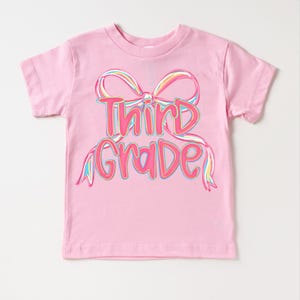 May include: A pink t-shirt with the words "Third Grade" in a colorful, bow-shaped design. The text is outlined in pink and filled with a gradient of colors, including yellow, blue, and green.