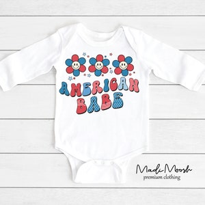 May include: White long-sleeved baby bodysuit with a retro design. The bodysuit features three smiling flower graphics in red and blue, with the words "AMERICAN BABE" in a matching font. The design is patriotic and playful.