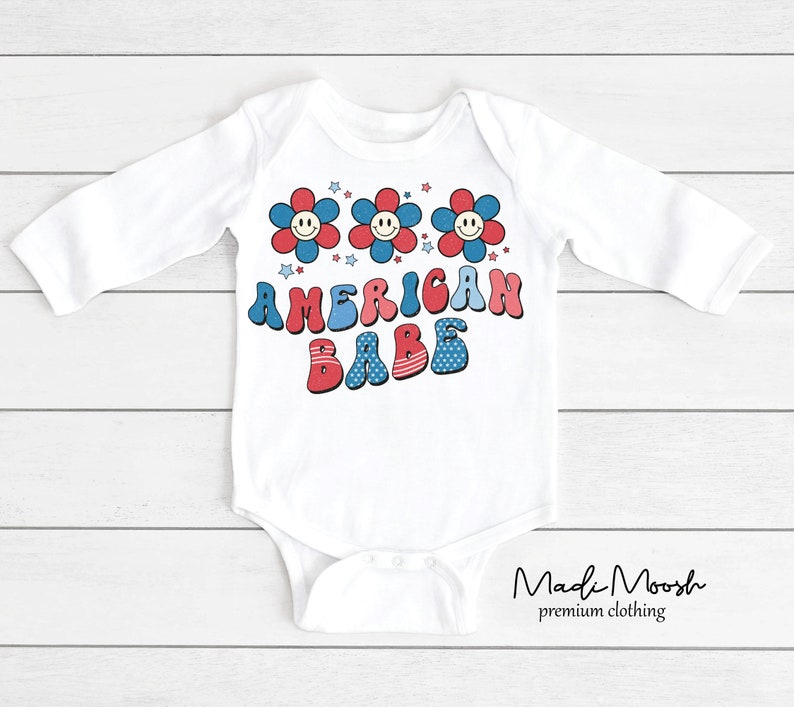 May include: White long-sleeved baby bodysuit with a retro design. The bodysuit features three smiling flower graphics in red and blue, with the words "AMERICAN BABE" in a matching font. The design is patriotic and playful.