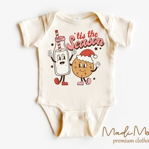 May include: A cream-coloured baby bodysuit featuring a cartoon milk carton and cookie, both smiling and giving peace signs. The text "'tis the season" is above them. The bodysuit is from Madi Moosh premium clothing.