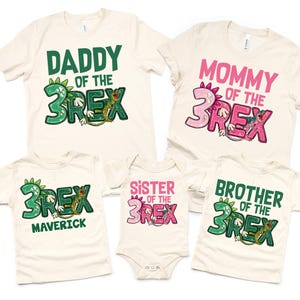 May include: Set of five cream-colored t-shirts with green and pink dinosaur graphics. The shirts say "Daddy of the 3Rex", "Mommy of the 3Rex", "Sister of the 3Rex", "Brother of the 3Rex", and "3Rex Maverick".