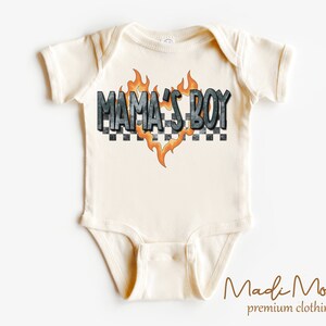 May include: Cream-colored baby onesie with the text "MAMA'S BOY" in a checkered and flame design. The onesie is short-sleeved and has a simple, classic design, perfect for a baby.