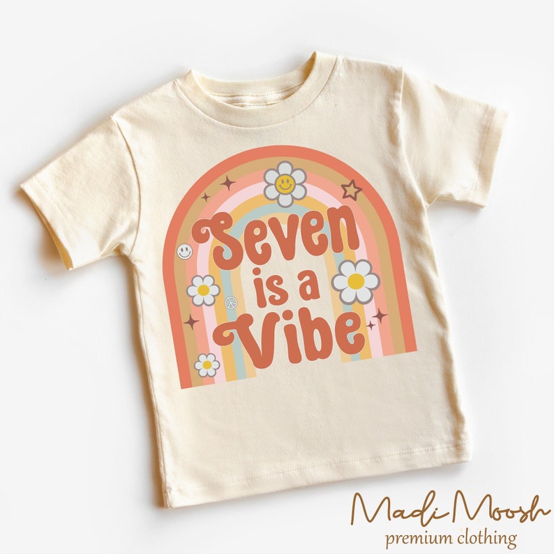May include: Cream-colored short-sleeved t-shirt featuring a retro rainbow design with the text "Seven is a Vibe." The design includes daisies, stars, and a smiley face. The brand name "Madi Moosh" is printed at the bottom.