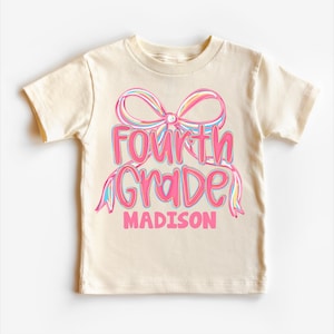 May include: A cream-colored t-shirt with a colorful bow and the words "Fourth Grade MADISON" in pink and blue. The shirt is made of soft material and is suitable for children.