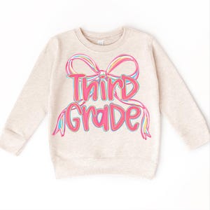 May include: A beige sweatshirt with the words "Third Grade" in pink, outlined in blue, with a rainbow-colored bow design. The sweatshirt has long sleeves and a crew neck, suitable for children.