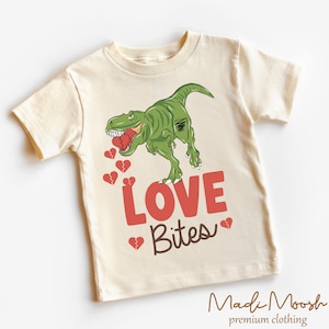 May include: Cream-colored t-shirt featuring a green dinosaur with a red heart in its mouth and broken hearts. The text "LOVE Bites" is printed in red and brown. The brand name "Madi Moosh" is at the bottom.