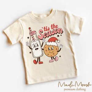 May include: A cream-coloured t-shirt with a cartoon illustration of a milk bottle and a cookie, both with arms and legs, giving peace signs. The text "'tis the Season" is above them. The brand name "Madi Moosh" is at the bottom.