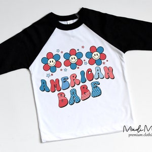May include: White baseball style t-shirt with black sleeves. The shirt features a retro design with three smiling flower graphics in red, white, and blue, and the words "AMERICAN BABE" in a matching font.