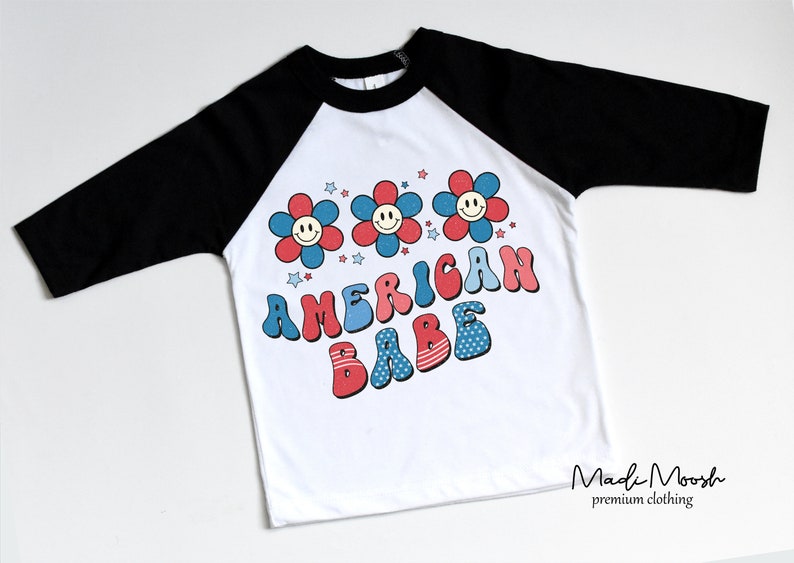 May include: White baseball style t-shirt with black sleeves. The shirt features a retro design with three smiling flower graphics in red, white, and blue, and the words "AMERICAN BABE" in a matching font.