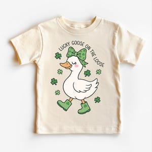 May include: Cream-colored t-shirt featuring a cartoon goose wearing a green bow, boots, and surrounded by shamrocks. The text "LUCKY GOOSE ON THE LOOSE" is printed above the goose in black.