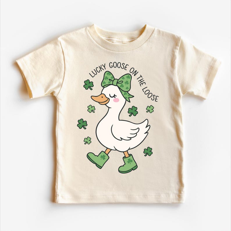 May include: Cream-colored t-shirt featuring a cartoon goose wearing a green bow, boots, and surrounded by shamrocks. The text "LUCKY GOOSE ON THE LOOSE" is printed above the goose in black.