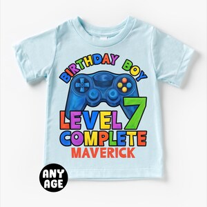 May include: Light blue t-shirt with a colorful graphic of a video game controller and the text "Birthday Boy Level 7 Complete Maverick".