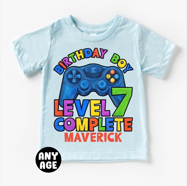 May include: Light blue t-shirt with a colorful graphic of a video game controller and the text "Birthday Boy Level 7 Complete Maverick".
